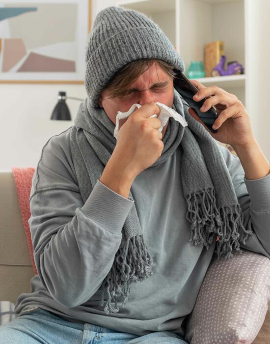 Sick young man with scarf around neck wearing winter hat wipes his nose talking phone sitting couch living room 1